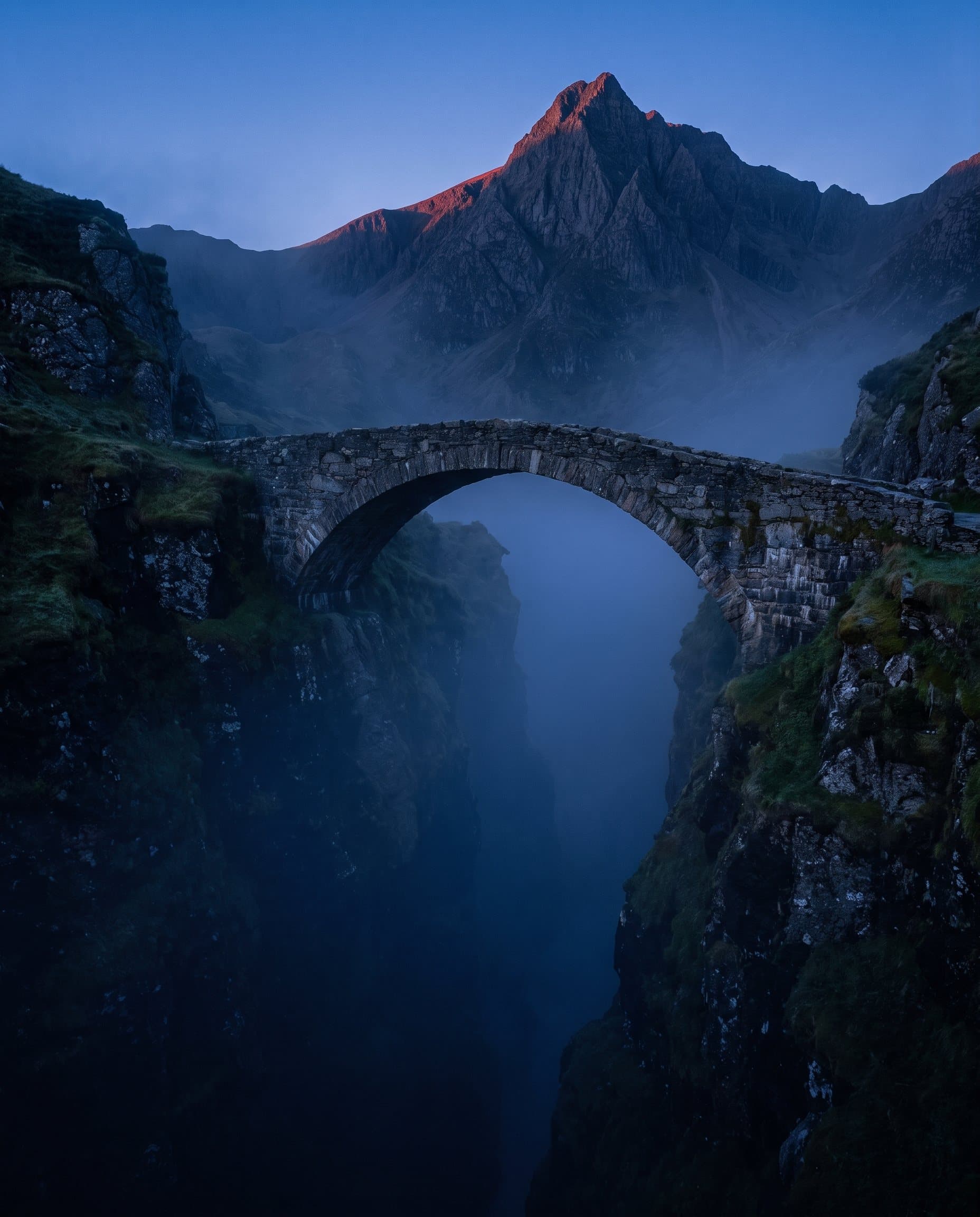 Steinbogenbrücke über eine tiefe Bergschlucht in der blauen Stunde, dahinter ein Gipfel mit rotem Morgenlicht am Grat