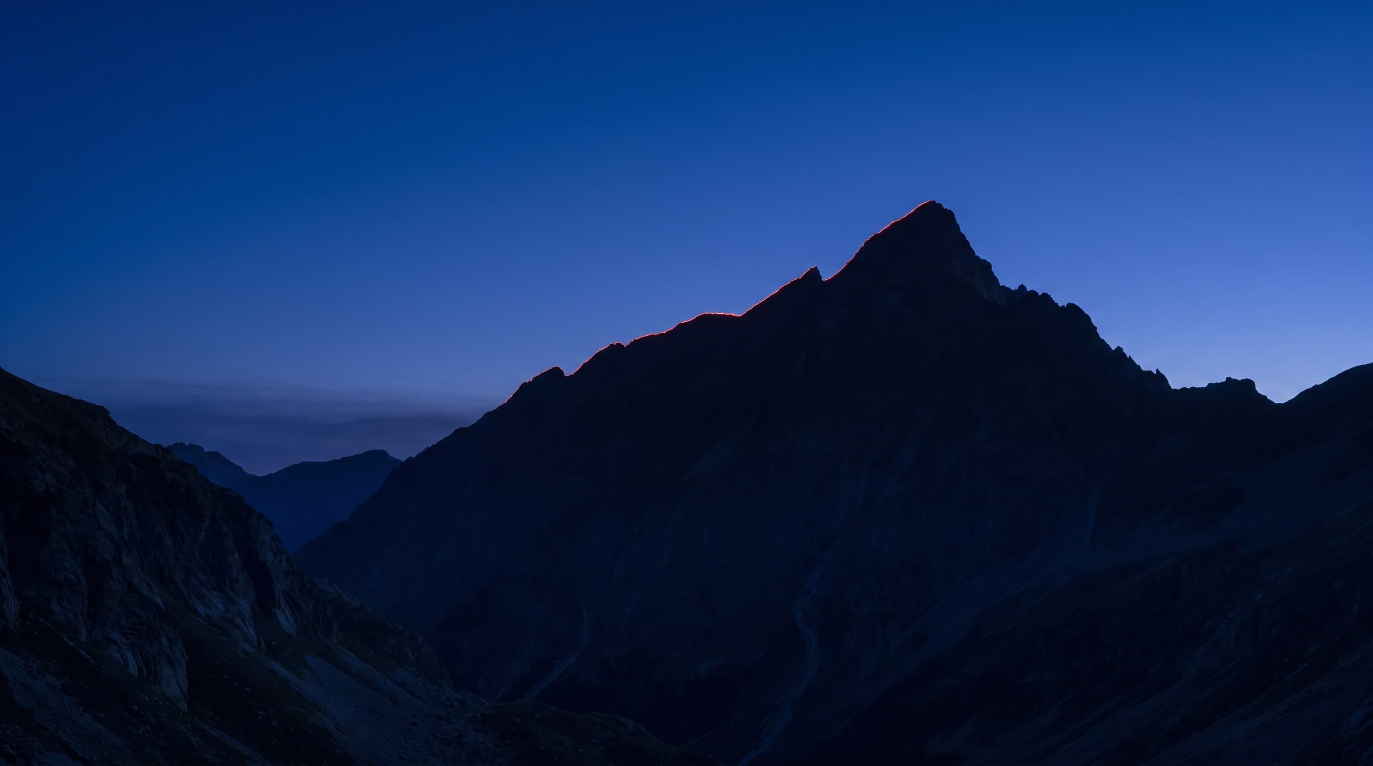 Spitzer Berggipfel in der blauen Stunde, der Grat mit einem roten Saum aus erstem Licht