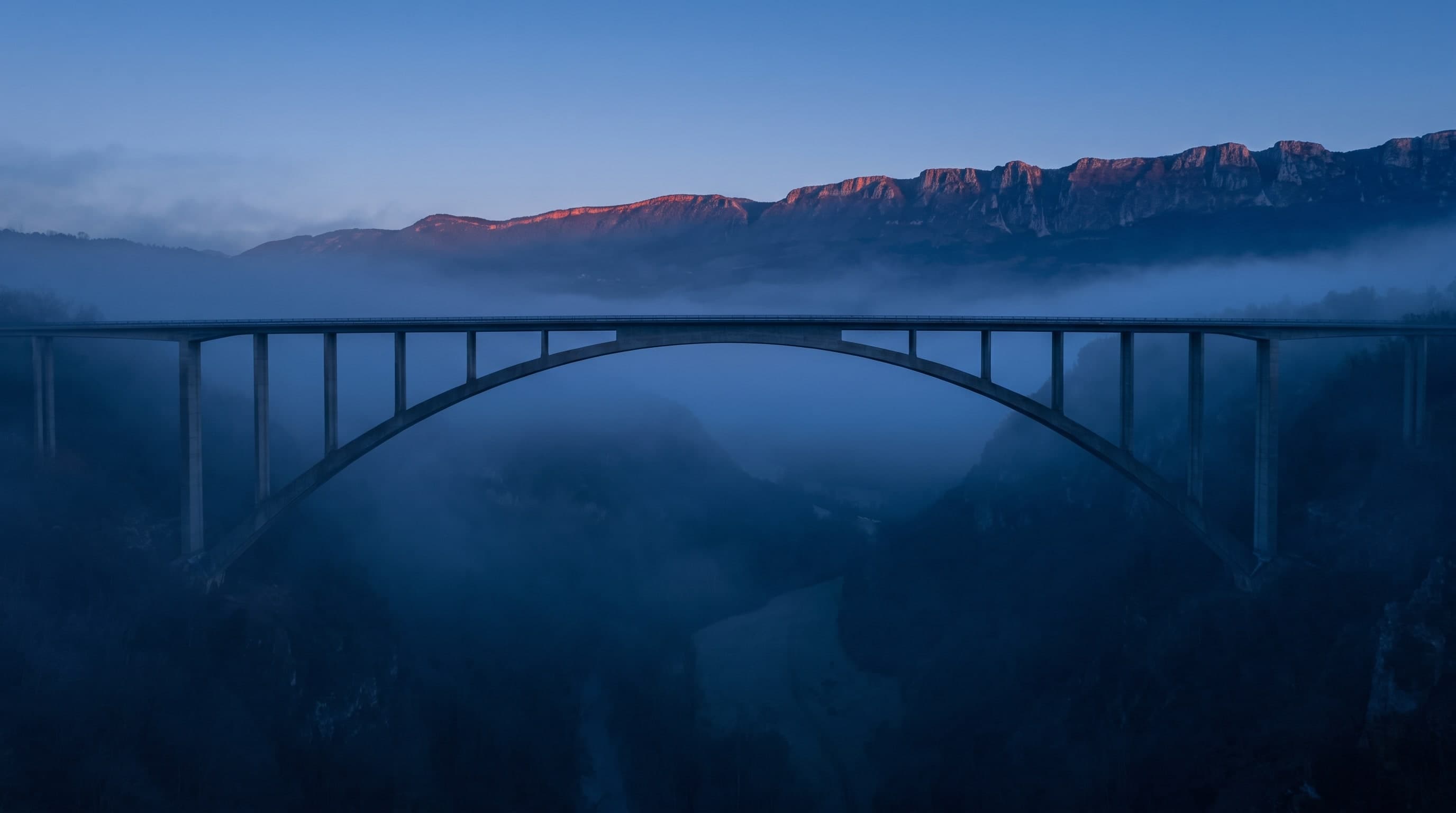 Schlanke Bogenbrücke über ein nebliges Tal, dahinter ein Bergkamm im ersten Morgenlicht