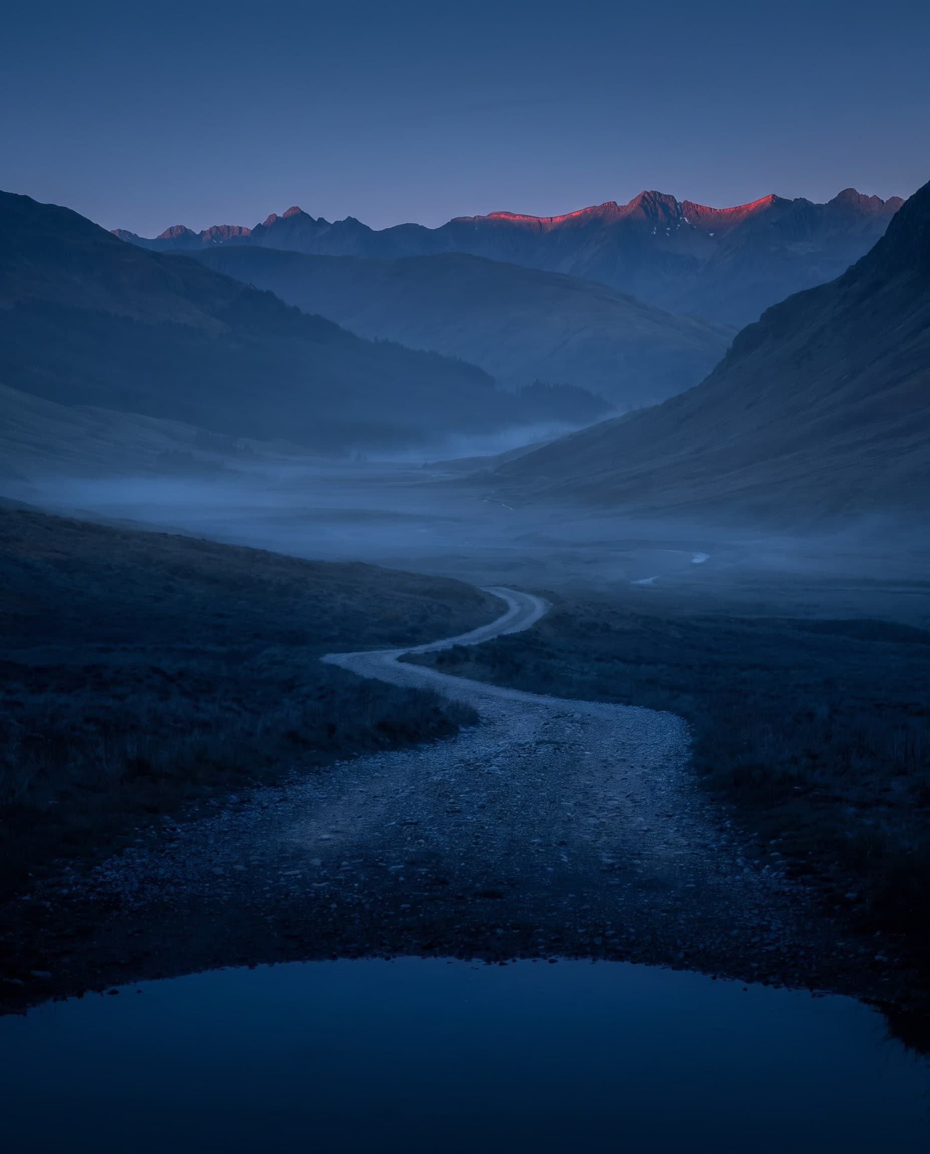 Schotterweg durch ein nebliges Bergtal, am Horizont Gipfel im ersten Licht